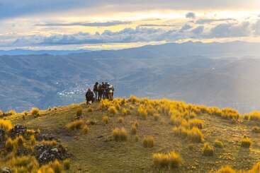 Un groupe de randonneurs se tient au sommet d'une colline verdoyante au coucher du soleil, entouré de touffes dorées et surplombant de vastes chaînes de montagnes sous un ciel spectaculaire rempli de nuages.