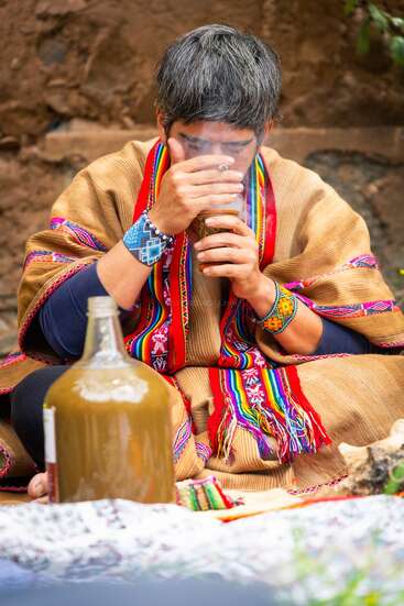 Une personne vêtue d'un costume traditionnel andin est assise en tailleur et boit dans une tasse. On aperçoit des vêtements colorés et des broderies de perles, ainsi qu'une grande cruche brune à proximité, suggérant un rituel.