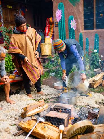 Trois personnes sont réunies autour d'un feu en plein air, préparant ou servant une boisson. Elles portent des vêtements traditionnels andins colorés. Des cactus peints aux couleurs vives décorent les murs rustiques environnants.