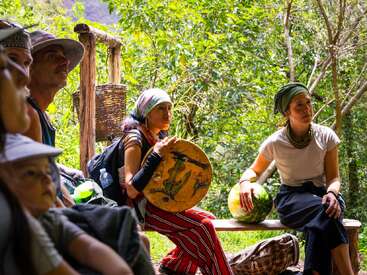 Un groupe de personnes est assis en plein air, entouré de verdure. Une femme tient un tambour peint, une autre pose sa main sur une pastèque. Elles semblent écouter attentivement.