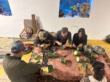 Un groupe de personnes est assis autour d'une table, chacune coupant des végétaux verts, peut-être des cactus, sur des planches à découper. La scène semble communautaire et culturellement riche, à l'intérieur.