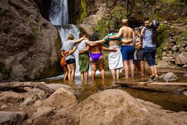 A group of people stands together in a circle, arms around each other, facing a beautiful waterfall in a rocky natural setting, enjoying nature and friendship.