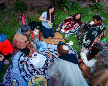 A group of people sit outdoors on colorful blankets, sharing food and drinks. Some wear traditional clothing, while others interact, smiling and enjoying each other's company.