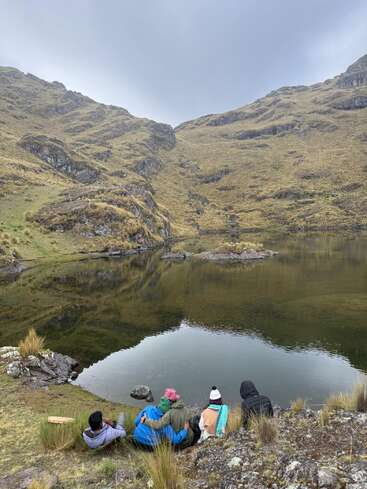 Four people sit by a serene mountain lake, surrounded by grassy hills and rocky terrain. The cloudy sky creates a calm, peaceful atmosphere in nature.