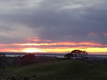 Das Bild zeigt eine ruhige Landschaft mit einem grasbewachsenen Hügel, Bäumen und einem Gewässer vor einem leuchtenden Sonnenuntergangshimmel mit rosa und gelben Farbtönen.