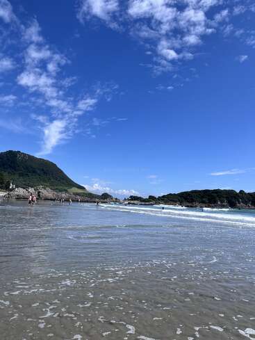 A beautiful beach scene with clear blue sky, gentle waves, people enjoying the water, distant green hills, and scattered clouds creating a peaceful, tropical atmosphere.