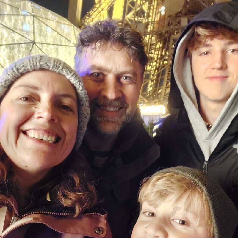 The image shows a family of four posing for a selfie in front of a large, lit-up structure, likely at night. The family is dressed warmly.
