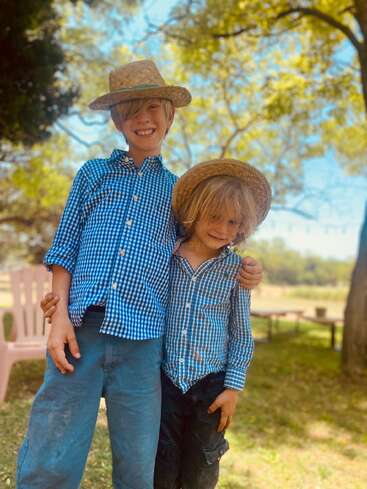 Dois meninos, vestindo camisas xadrez azuis e chapéus de palha combinando, estão sorridentes de braços dados ao ar livre, sob as árvores, desfrutando de um dia ensolarado e despreocupado.