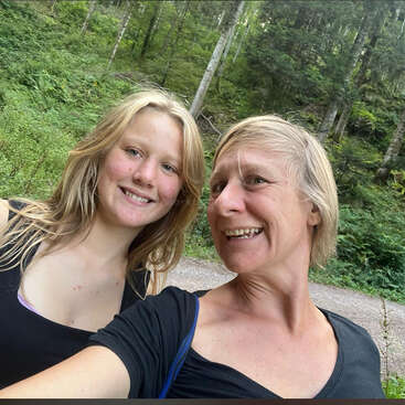 Duas mulheres sorridentes tiram uma selfie alegre juntas ao ar livre, cercadas por árvores verdes exuberantes e um caminho de terra, capturando um momento alegre durante sua caminhada na natureza.