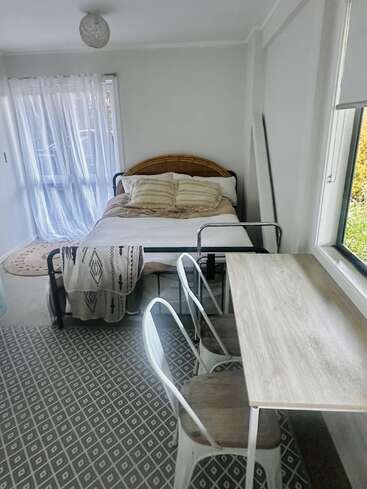Un dormitorio acogedor y minimalista con una cama con cojines a rayas, una manta ligera, un pequeño escritorio con dos sillas, una alfombra estampada y cortinas transparentes que dejan pasar la luz natural.