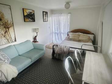 Se trata de un dormitorio acogedor y luminoso que cuenta con una cama, un sofá azul claro, un escritorio con dos sillas, un gran ventanal con cortinas transparentes, cuadros en las paredes y una alfombra estampada.