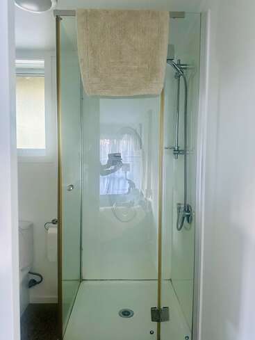 En esta imagen se ve una mampara de ducha moderna con puertas de cristal y un cabezal de ducha de mano. Sobre el marco superior de la puerta hay una toalla de color beige claro.
