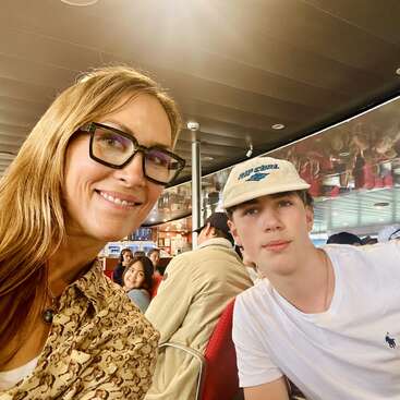 A woman in glasses and a young man in a hat smile for a selfie on public transport, with other passengers and modern interior visible in the background.