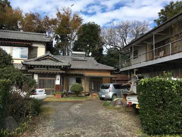 A imagem mostra uma casa serena em estilo japonês com um telhado de telhas cinza, cercada por uma vegetação exuberante e uma entrada de automóveis de cascalho, exalando uma sensação de tranquilidade e beleza natural.