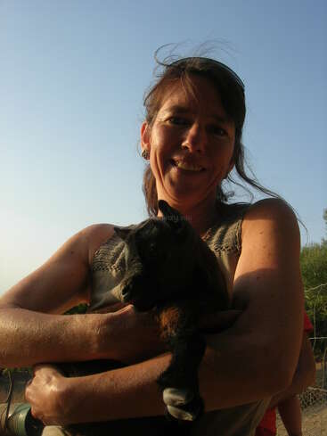 Une femme souriante est à l'extérieur, sous un ciel clair, tenant une petite chèvre noire dans ses bras, profitant de la lumière chaude du soleil et de l'environnement naturel.