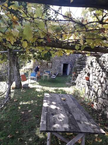 Una casa rústica de piedra con una pérgola cubierta de enredaderas da sombra a una vieja mesa de madera. Una persona está de pie cerca de unas sillas azules, rodeadas de vegetación, creando un ambiente tranquilo.