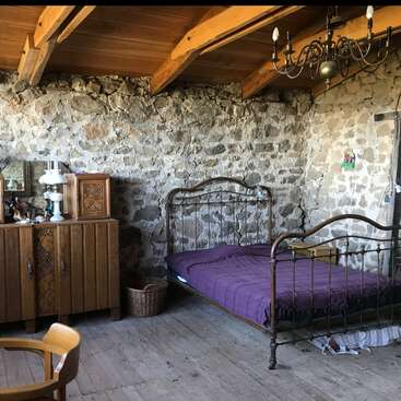 Este dormitorio rústico presenta una pared de piedra, vigas de madera, una cama vintage de metal con una manta morada, muebles de madera, lámpara de araña, cesta de mimbre y suelos de madera.