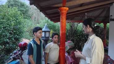 A woman in a white shirt is talking to two boys outside a house. The boys appear attentive. There are motorbikes and greenery in the background.
