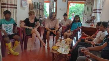 A diverse group of people sits together in a cozy living room, chatting and smiling. Refreshments and snacks are placed on a small table in the center.