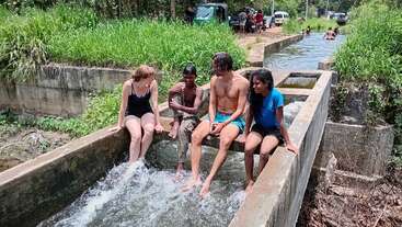 Four people sit on a concrete water channel, feet in running water, enjoying a sunny day. Lush greenery and more people are visible in the background.