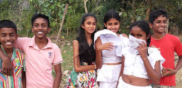 Three young girls pose outdoors, smiling and playful. One girl covers her face with a white cloth. Lush green trees provide a natural background for them.