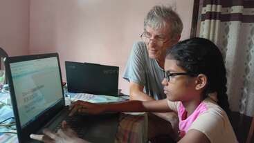 A young girl with glasses is typing on a laptop while an older man, also with glasses, sits beside her, supervising and providing guidance in a cozy room.