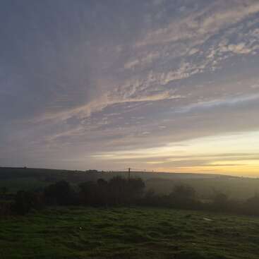 A serene landscape at dusk features a grassy field, distant hills, and soft, gently layered clouds in the sky illuminated by the setting sun's golden glow.