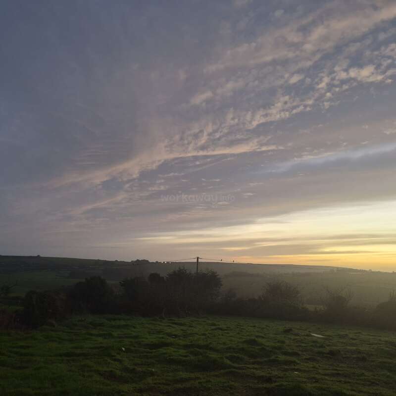 Uma paisagem serena ao entardecer apresenta um campo gramado, colinas distantes e nuvens suaves e delicadamente dispostas em camadas no céu, iluminadas pelo brilho dourado do sol poente.
