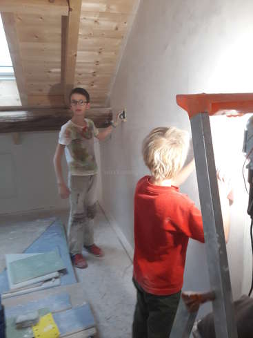 The image depicts two young boys engaged in painting a room, with one standing on the left and the other on the right, both holding paint rollers.