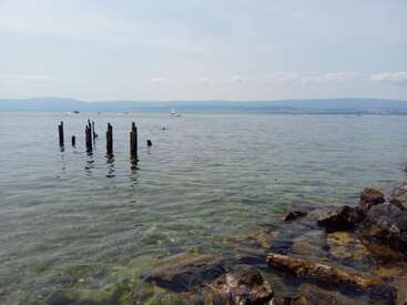 The image depicts a serene lake scene with a rocky shoreline, featuring a few wooden posts protruding from the water, set against a hazy blue sky.