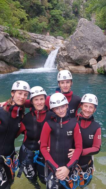 Five people in wetsuits and helmets stand in front of a natural pool and waterfall, surrounded by rocks and lush green forest, enjoying outdoor adventure.