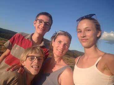 A group of four people, smiling and happy, take a selfie outside on a sunny day with blue skies and hills in the background. Natural, joyful moment.