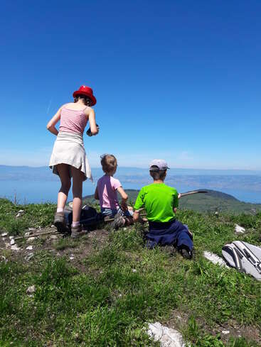 L'image représente trois enfants assis sur une colline herbeuse, surplombant une vaste étendue d'eau et des montagnes lointaines sous un ciel bleu clair.
