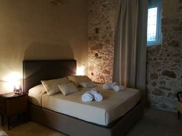 Das Bild zeigt ein ruhiges Schlafzimmer mit Bett, Nachttisch und Fenster, das mit seiner Steinwand und der beigen Farbgebung ein Gefühl von Ruhe und Entspannung vermittelt.