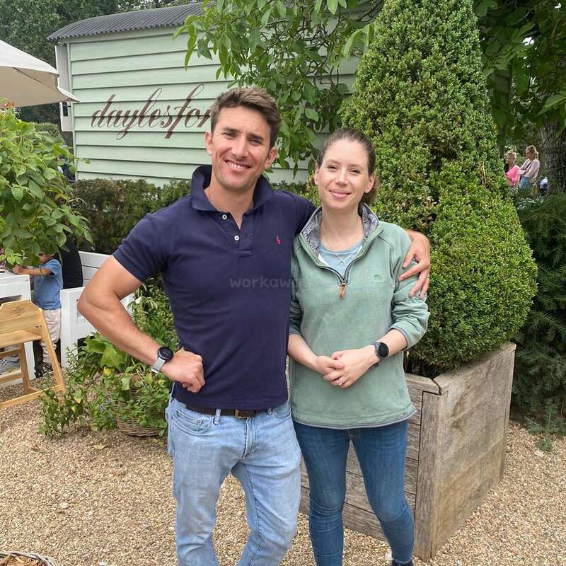 A smiling man and woman pose together outside, surrounded by greenery. The background shows a sign reading "daylesfo," wooden siding, and relaxed outdoor atmosphere. Both wear casual clothes.