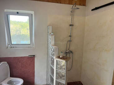 Esta imagen muestra un cuarto de baño sencillo con un inodoro, una ventana y una ducha a ras de suelo. La ducha cuenta con un separador de bloques de vidrio y un cabezal de ducha moderno.