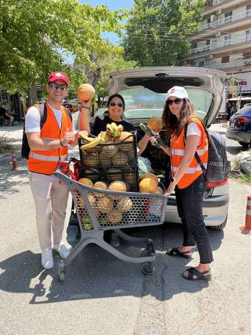 Three people wearing orange safety vests and hats are smiling beside a shopping cart full of melons, next to an open car trunk, outdoors on a sunny day.