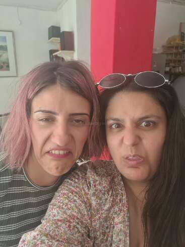 Two women are making funny, exaggerated grimacing faces for a selfie. One has pink hair, the other wears sunglasses on her head. Bright, casual indoor setting.