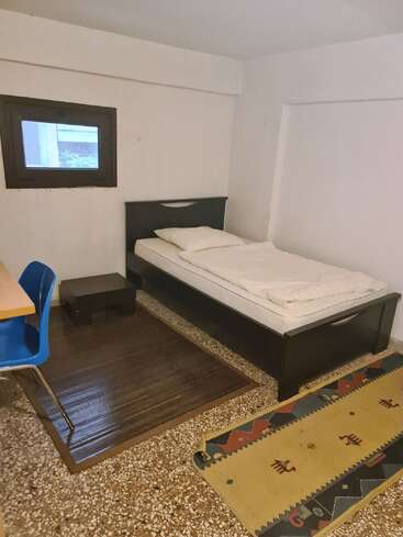 This image shows a simple, minimalist bedroom with a single bed, pillow, white bedding, a small black nightstand, blue chair, desk, and two rugs.