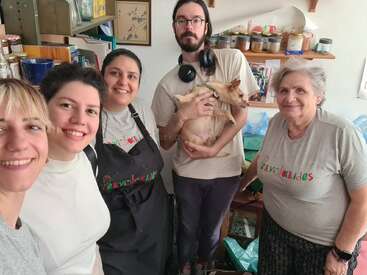 Five people, three women in matching shirts, one man holding a small dog, and an elderly woman, smiling together in a cozy, cluttered room.