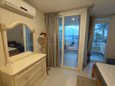 La imagen muestra un sereno dormitorio con un gran espejo, una cómoda y una puerta corredera de cristal que da a un balcón con vistas a una tranquila masa de agua.