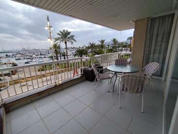 La imagen representa un sereno balcón con barandilla blanca, con vistas a un puerto deportivo, una mesa y sillas, palmeras y un cielo nublado.