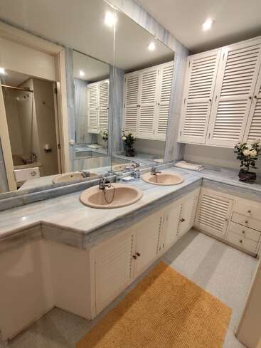 La imagen muestra un cuarto de baño con un gran espejo, dos lavabos y armarios blancos, con una encimera de mármol y una alfombra amarilla en el suelo.