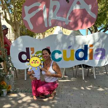 La imagen muestra a una mujer y un niño posando delante de un cartel con la palabra "FIRA" y letras de colores, frente a un camino de ladrillos y árboles.