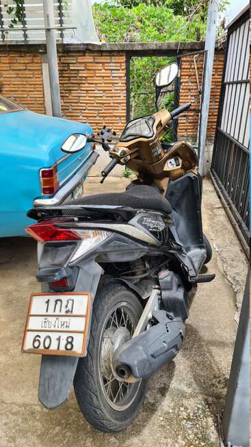 A black and gold scooter with a Thai license plate is parked next to a classic blue car in a small, walled courtyard with greenery.