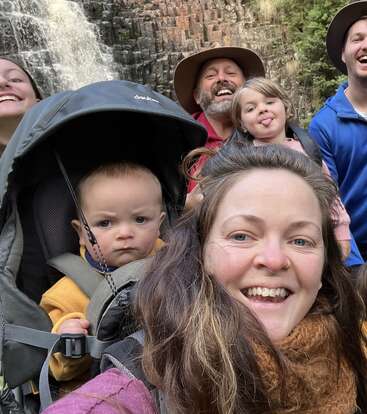 L'image représente une famille de six personnes posant pour un selfie devant une cascade, avec un bébé dans un porte-bébé et une femme au premier plan.