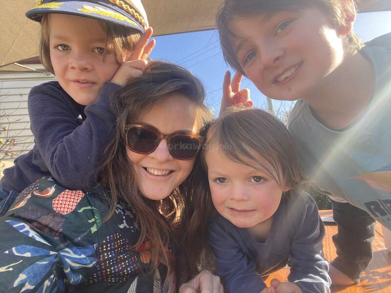 Une femme joyeuse portant des lunettes de soleil pose à l'extérieur avec trois enfants souriants, deux d'entre eux arborant des oreilles de lapin. La lumière du soleil et les expressions détendues et heureuses créent une atmosphère joyeuse.
