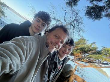 Three people smile for a selfie outdoors by a scenic lakeside. Bare trees, green branches, and calm blue water create a peaceful, natural background.