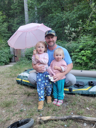 The image depicts a man sitting on an inflatable raft with two young girls, one holding a pink umbrella, in a wooded area with lush greenery and trees in the background.