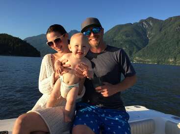 The image depicts a family of three on a boat, with the mother holding a baby and the father standing beside them, set against a scenic mountainous backdrop.